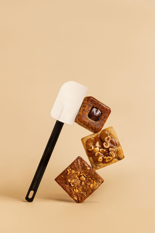 Carrés pâtissiers aux noisettes et chocolat posés sur une spatule, fond beige uni