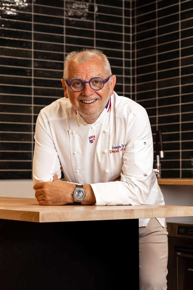 Chef en blouse blanche accoudé à un comptoir en bois clair, fond de carreaux noirs